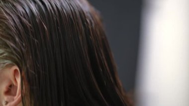 Live camera moves along brunette straight laminated hair as hairdresser spraying conditioner in slow motion. Unrecognizable young Caucasian woman doing keratin treatment in hair salon indoors