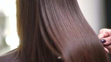 Close-up shaking long laminated hair with hair brush in slow motion. Brunette unrecognizable Caucasian young woman after keratin treatment in beauty parlor indoors with hairstylist
