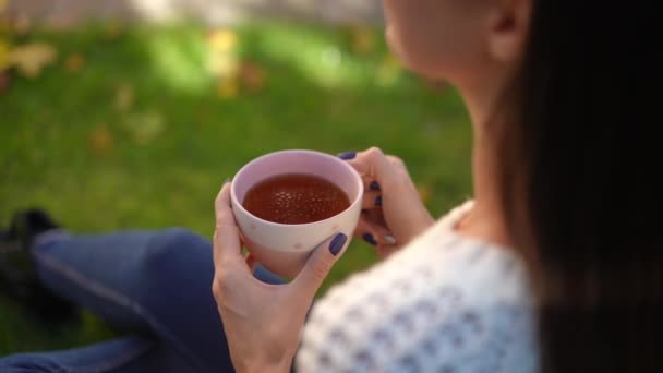 一个身材苗条的年轻女子坐在绿草上 一边喝着茶 一边拍拍肩膀 无法辨认的白人女士在后院享受户外热饮 — 图库视频影像