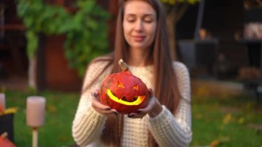 İçinde ışık olan Cadılar Bayramı balkabağı ve arka planda Jack-o-Lantern 'i yavaş çekimde geren bulanık beyaz kadın. Arka bahçede tatil süslemeleriyle tanınmayan mutlu genç bayan.