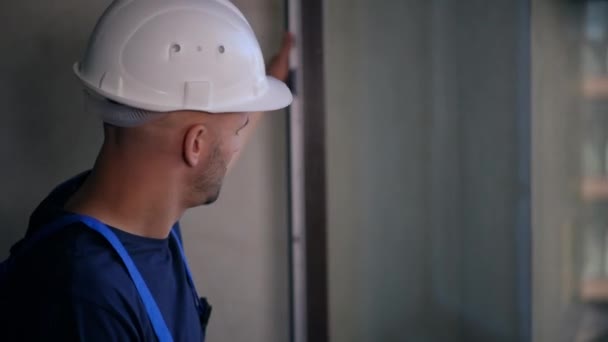 自信的建设者戴着硬礼帽 用新的扁平思维检查室内的窗户 身穿制服的白人青年男子的侧视图 规划公寓翻新 建筑和维修概念 — 图库视频影像