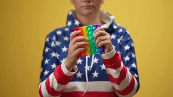 黄色の背景に立って捨てるカラフルな感覚玩具を絞る神経質な不安千年紀の女性 若い白人女性はアメリカ国旗色のパーカーにフィジェットトイで強調した 応力管理 — ストック動画