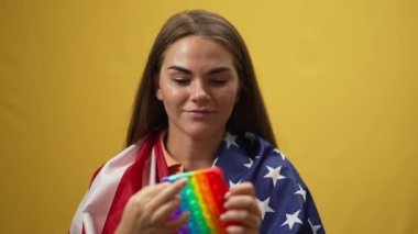 Gülümseyen büyüleyici kadın ağır çekimde oyuncağını oynuyor sarı arka planda Amerikan bayrağı omuzlarında duruyor. Moda oyuncaklarından hoşlanan mutlu, kendine güvenen bir kadının portresi