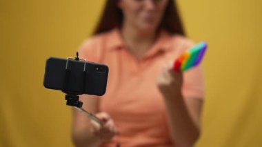 Akıllı telefonlu selfie çubuğu ve sarı arka planda hareketli oyuncaklar reklamındaki bulanık genç kadın. Kafkasyalı kendine güvenen blogcu sosyal medyada canlı yayında duyusal oyuncağa danışmanlık yapıyor
