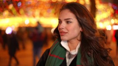 Çekici, pozitif esmer, kahverengi gözlü kadın Noel gecesi şehir merkezinde durmuş etrafa bakıyor ve gülümseyen kameraya bakıyor. Açık havada poz veren kendine güvenen beyaz bir kadının portresi.