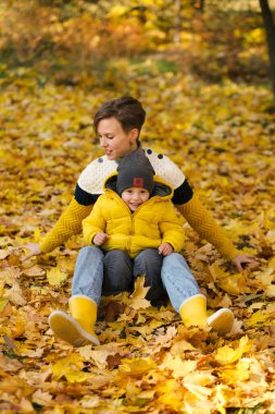 Küçük oğlunu kucaklayan ve sonbahar parkında sarı akçaağaç yapraklarının arasında oturan mutlu genç anne.