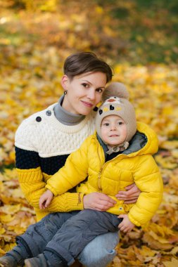 Anne ve oğlu sonbahar parkında sarı yaprakların arasında kameraya bakıyor.