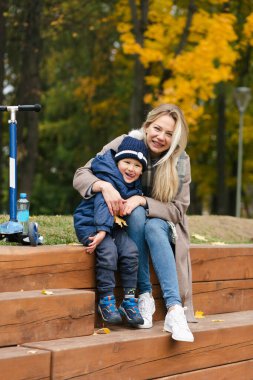 Anne oğlunu kucaklıyor, tahta parkta oturuyor ve gülümsüyor.