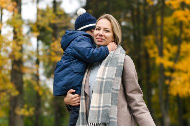 Mutlu anne oğlunu sonbahar parkında tutuyor ve kameraya bakıyor.