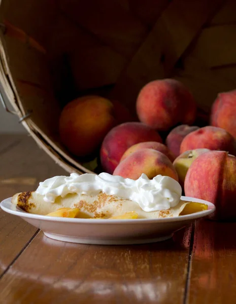 Warm crepe and peaches with whipped cream, sit in front of a basket of fresh peaches used to help make this decedent treat