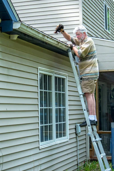 An older man carefully stands on a ladder and cleans leaves and debris from his home\'s gutters