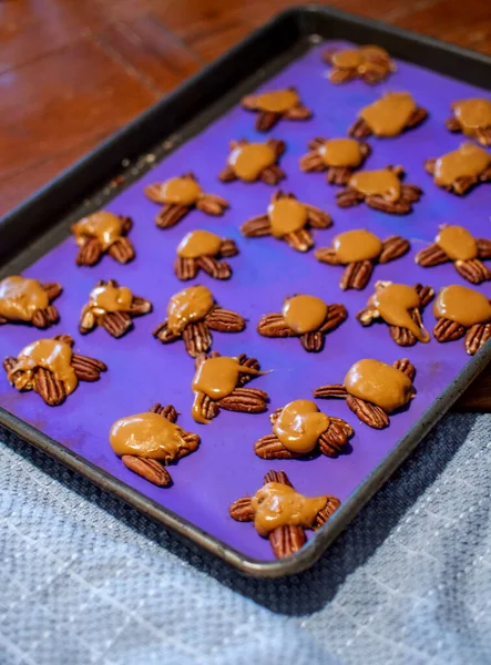 A cookie sheet covered by a silicone mat, holds a array of turtle candies made with caramel and pecans then topped with chocolate