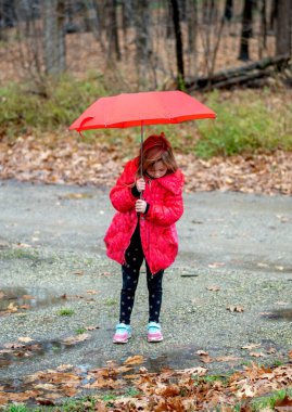Kırmızı paltolu genç kız, şemsiyesini tutuyor ve atlamak için su birikintisi arıyor.