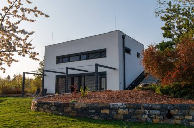Modern, beyaz, küp, zarif, minimalist tarzda pasif bir ev. Büyük panoramik pencereli, gün batımında sıcak bir bahçede korunan gri panjurlar. Tahta teras. Bahçe mobilyası.