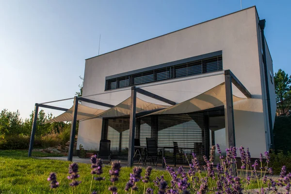 Modern, white, cube, elegant, minimalist style passive house with large panoramic windows, grey shutters in maintained garden. Wooden terrace. Sunny day. Lavender in front.