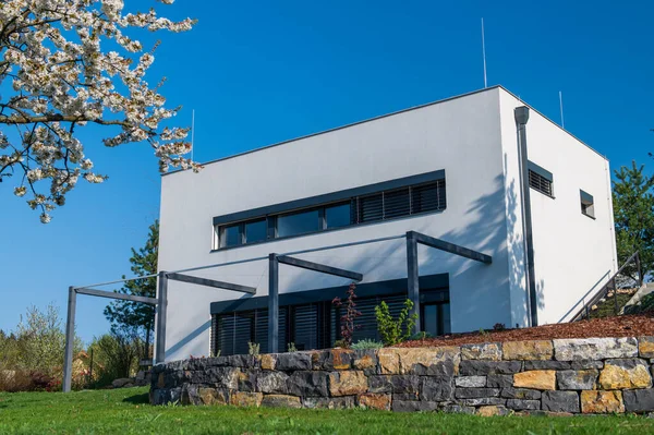 Modern, white, cube, elegant, minimalist style passive house with large panoramic windows, grey shutters in maintained garden. Wooden terrace. Sunny day.