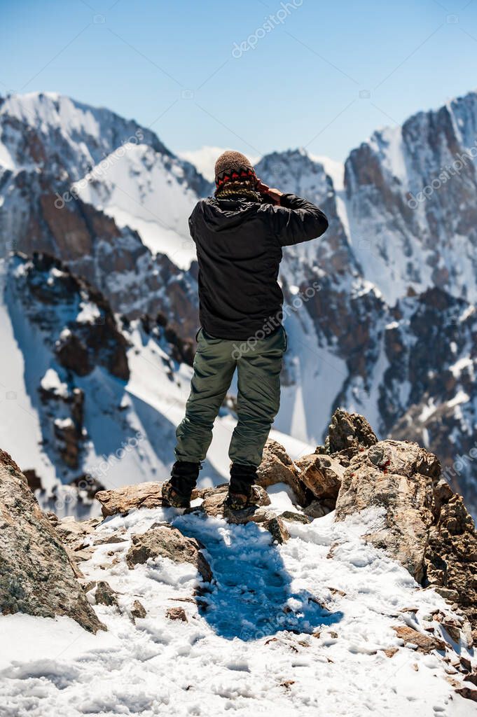 Young man trekker photographer taking pictures with camera on top of Pik Uchitel peak . Ala Archa Alpine National Park Landscape near Bishkek, Tian Shan Mountain Range, Kyrgyzstan, Central Asia.