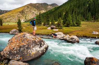 Genç adam ve kadın, Karakol vadisinde Ala Kul Gölü 'ne giden küçük tahta köprüden vahşi turkuaz nehri geçiyorlar. Karakol vadisi, Issyk-kul bölgesi, Ala-kul Gölü Terskey Alatau dağ sırası, Kırgızistan, Orta Asya.