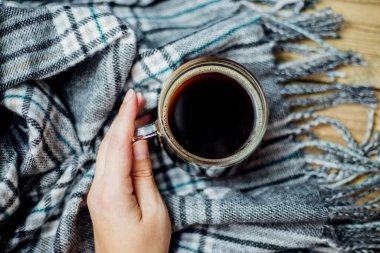 Bir kadının eli, cam bardağı sıcak kahveyle tutuyor. Hafif ahşap zemin üzerinde püsküllü sıcak bir ekose eşarp var. Konfor ve sıcaklık kavramı.