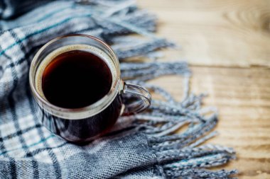 Hafif ahşap zemin üzerinde püsküllü sıcak bir ekose eşarp var. Sade kahveli cam bardak yumuşak gri kumaşın üzerinde duruyor. Konfor ve sıcaklık kavramı.