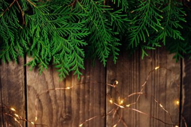 Yeşil thuja dallarının Noel ışıklarının arka planı. Ahşap tahtaların kahverengi yüzeyinde şenlikli parlak bir çelenk dallarla sarmalanmıştır. Noel ağacı süslemeleri konsepti.