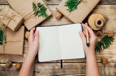 Kadın elleri kalemle deftere yazar. Tahta bir arka planda Kraft kağıdında kutular var. Çevre dostu materyal. El yapımı sanat eseri. Noel ve Yeni Yıl kavramı.