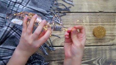 Women's hands hold a glass bottle with a garland of Christmas lights. Decoration made with your own hands.Warm grey scarf or plaid on a wooden background