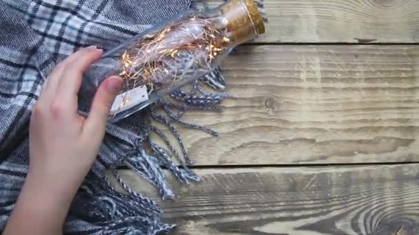 女性の手はクリスマスライトのガーランドとガラスボトルを保持します 暖かい灰色のスカーフや木製の背景に再生 — ストック動画
