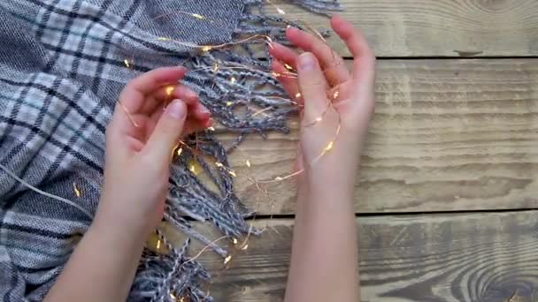 女性の手はクリスマスライトのガーランドを保持暖かい灰色のスカーフや木製の背景に再生 — ストック動画