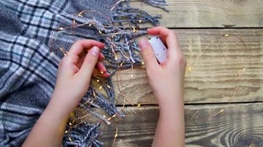 Women's hands hold a Christmas garland. Lights on and off. Grey scarf or plaid on a wooden background.