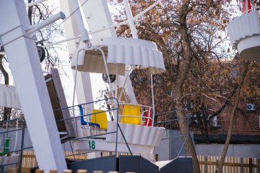 The white cab of the Ferris wheel against the background of trees. A public amusement park. Attractions for children and adults.