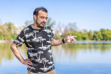 Gölde sakallı, olgun bir adam kameraya bakıyor. Bir kolu akimbo, diğeri de yan tarafı işaret ediyor. Boşluğu kopyala