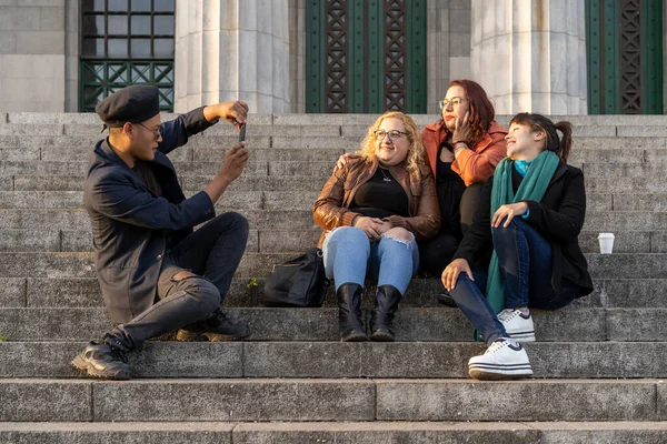 Çok ırklı bir arkadaş grubu merdivenlerde oturmuş sohbet ediyor, gülüyor ve fotoğraf çekiyor.