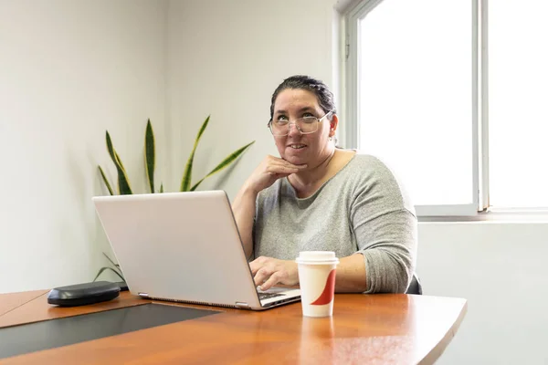 Mature business woman sitting at head of table in office meeting room, working on her computer, looking at someone standing out of frame