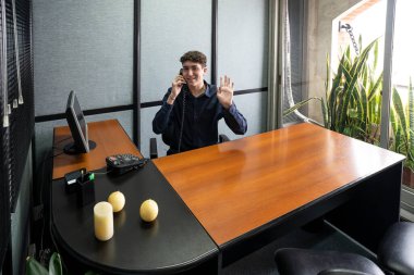 Businessman sitting at his desk talking on the phone looking at the camera and waving