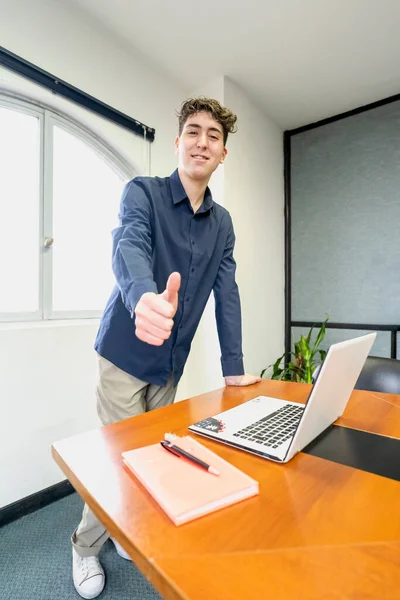 Young businessman standing at the head of the meeting table, smiling and looking at the camera with his thumb up