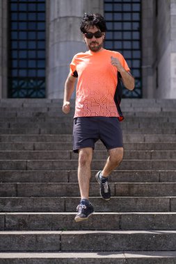 Yakışıklı, olgun, sakallı, atletik, 40 yaşlarında, turuncu tişörtlü, merdivenlerde koşan bir adamın portresi..