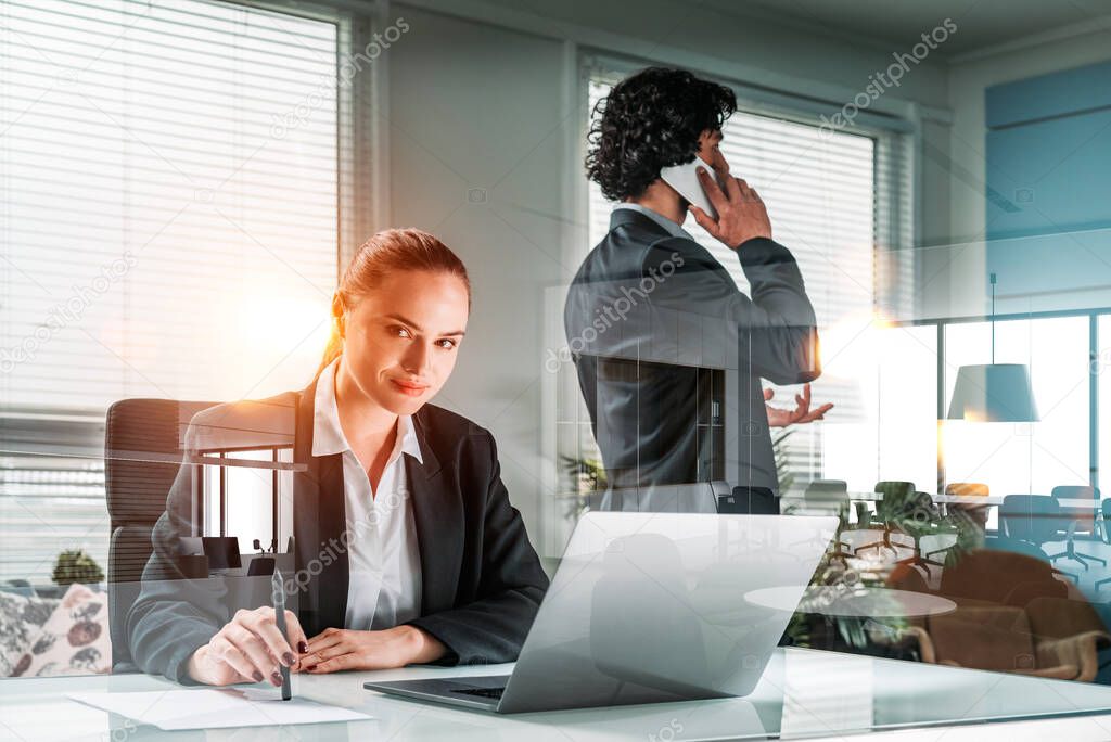 Businessman and businesswoman wearing formal suits work together talking on smartphone and using laptop. Office workplace in the background. Concept of teamwork and cooperation