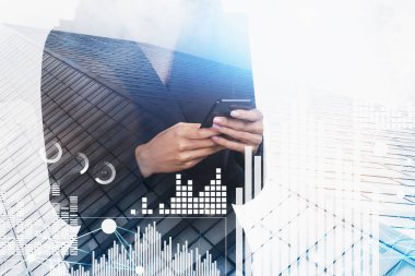 Akıllı telefonları olan bir iş kadını başarılı bir ticaret yapmak için ekrana dokunuyor. Borsa davranışı kavramı. Hologram finansal grafikleri. Bulanık alçak açılı gökdelenler cephe.
