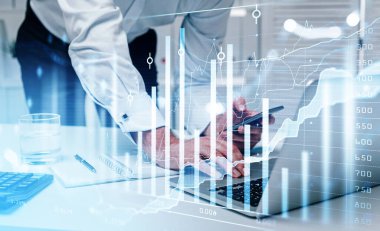 Beyaz tişört giyen bir iş adamı dizüstü bilgisayarına mesaj yazıyor. Arkasında akıllı telefon olan bir ofis işyeri. Finansal grafik ve grafik, önplanda çubuk diyagramı