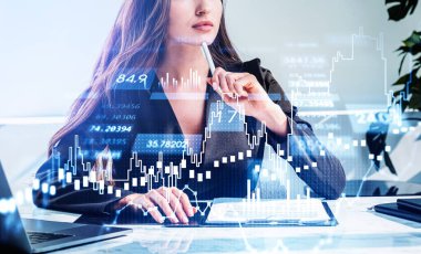 Resmi takım elbise giyen iş kadını dizüstü bilgisayarın önünde not alıyor. Arka planda ofis işyeri var. Finansal tablolar ve grafikler, ön planda bir şamdan. Borsada ticaret kavramı