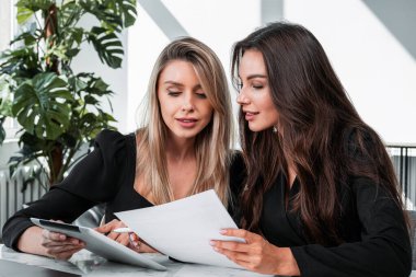 Siyahlı iki ofis kadını tablette, iş odasında fabrika ve iş belgeleriyle birlikte çalışıyorlar. Takım çalışması ve işbirliği kavramı