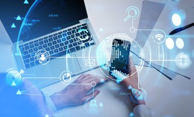 Beyaz gömlekli bir iş adamı akıllı telefondan mesaj yazıyor. Arkasında dizüstü bilgisayar olan bir ofis işyeri. Önplandaki sanal kürenin hologramıyla dijital arayüz. İş kavramı
