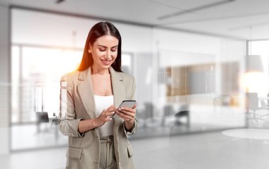 Ofisteki bej takım elbiseli kadın konferans odasının arka planında mobilyalı telefon kullanıyor. İşçi ve teknoloji kavramı