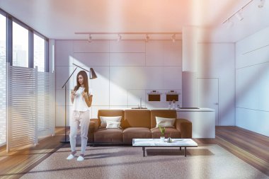 Telefon kullanan, evin içinde kahve içen, parke zemin ve halı üzerinde kahverengi kanepe olan genç bir kadın. Mutfak rafı ve masa, şehir manzarası ve beyaz ekran