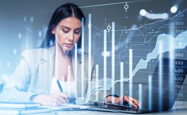 Resmi takım elbise giyen iş kadını dizüstü bilgisayarda çalışıyor ve not alıyor. Arka planda ofis işyeri var. Finansal grafik ve bar diyagramı. Başarılı iş ticareti kavramı