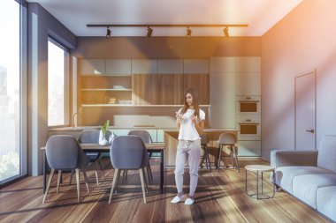 Genç bir kadın telefon kullanıyor, içeride kahve içiyor. Gri koltuklar ve kanepe, New York manzaralı ön manzara. Mutfak seti ve minimalist mobilyalar, mercek fişeği