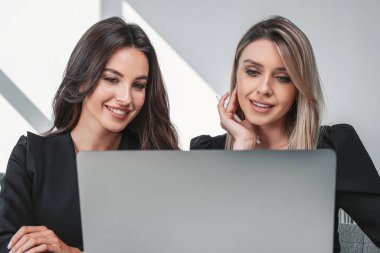 Resmi takım elbiseli iki gülümseyen iş kadını dizüstü bilgisayarda birlikte yazıyorlar. Arka planda masa olan ofis işyeri. Çalışma süreci ve takım çalışması kavramı
