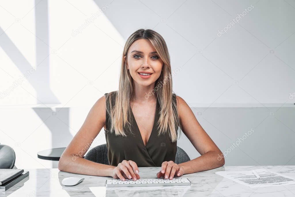 Office woman smiling, looking at the camera with hands on keyboard. Desk with papers and mouse. Manager working in office room. Concept of secretary and reception