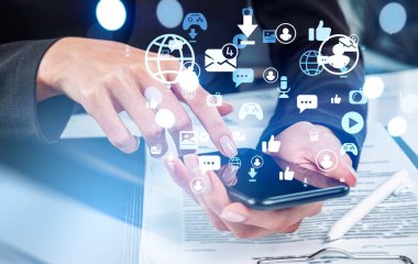 Resmi takım elbiseli iş kadını akıllı telefondan sosyal medyada geziniyor. Bilgisayarlı ve arka planda not defterleri olan ofis işyeri. Sanal kürenin simgeleri, ileti, oynat, kamera, mikrofon